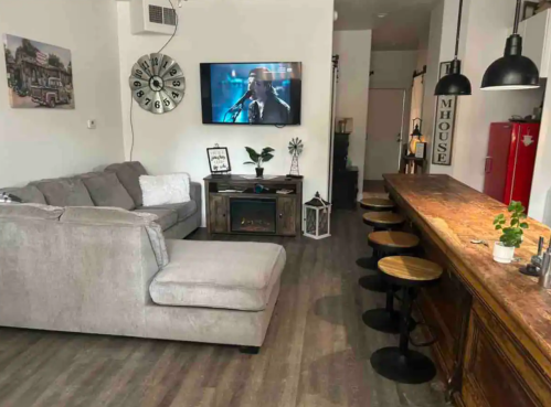 Cozy living room with a gray couch, wooden bar, and a TV displaying a performance. Modern decor and warm lighting.